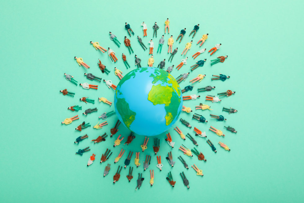 Kleine blaue Kugel, die mit den Kontinenten bemalt ist und mit vielen kleinen Menschen als Figuren, die im Kreis in mehreren Reihen um die Weltkugel gelegt sind